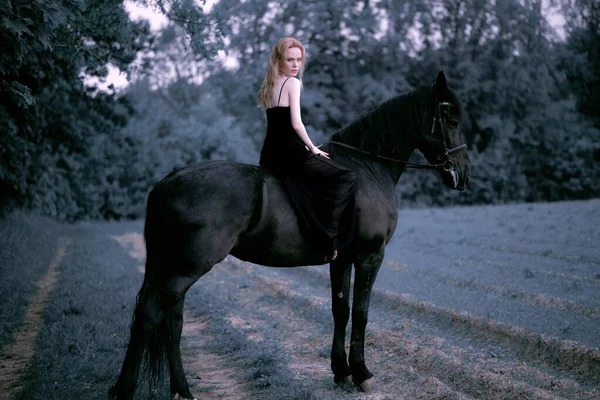 美しい若いブロンドの女性は黒いドレスを着て 黒い馬に乗って 暗い悲観的でゴシックな雰囲気 — ストック写真
