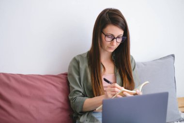 Konsantre genç kadın serbest çalışan notlar alıyor. Evden çalış..