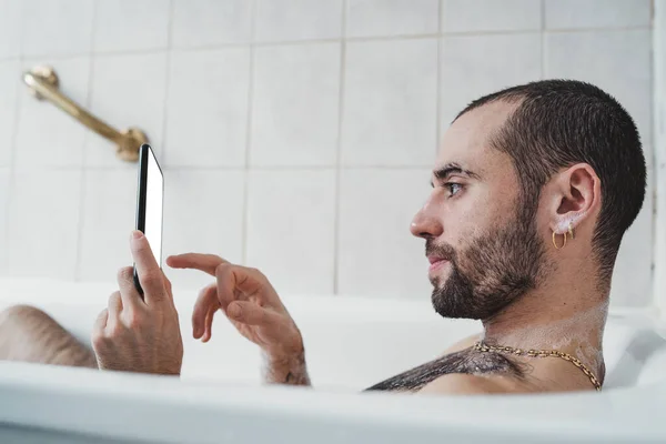 白人男子在浴缸里放松地看着白色的手机屏幕 — 图库照片