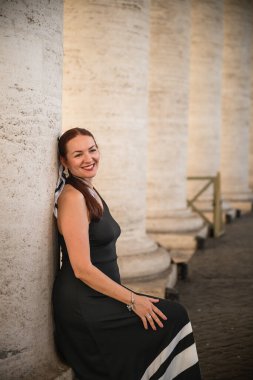 Siyah elbiseli kadın güneş ışığında antik sütunların yanında duruyor.