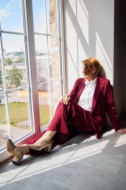 Kırmızı elbiseli kadın, parlak modern mekanda pencere kenarında dinleniyor.