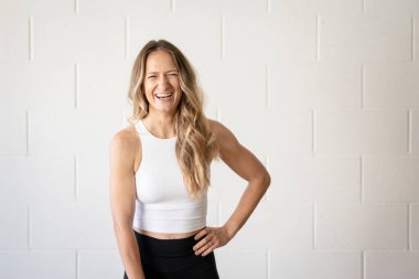 Beyaz duvarın karşısında spor kıyafetli, gülümseyen kadın.