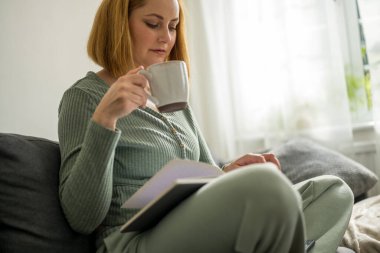Orta yaşlı bir kadın bir fincan kahveden ve defterden kitap okumaktan zevk alıyor.