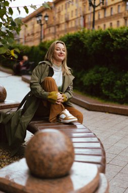 Gülümseyen kadın sıradan bir sonbahar modasında bankta oturuyor, yeşil ceketli.