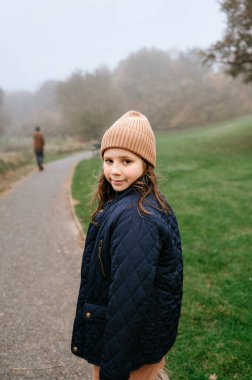 Genç bir kız sisli bir yolda arkasında yürüyen biriyle gülümsüyor..