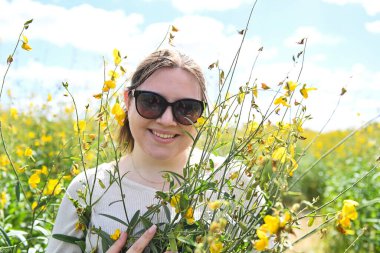 Uzun sarı çiçeklerin arasında, güneş gözlüklü, gülümseyen kadın..