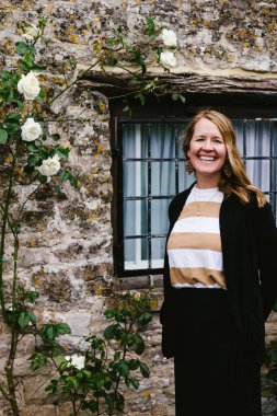 Sarışın kadın taş duvar, pencere ve gül çalısıyla gülümsüyor.