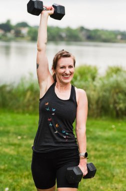 Gülümseyen kadın gölün kenarındaki dambılı kaldırıyor.