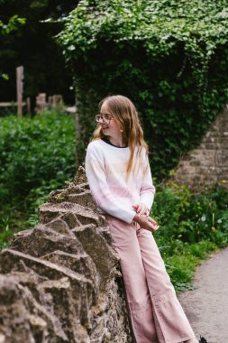 Sarışın genç kız Cotswold köyündeki antik taş duvara yaslanıyor.