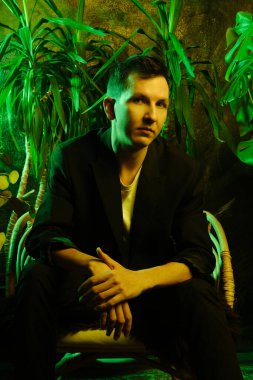 Canlı yeşil bitkilerin arasında oturmakta olan şık bir adam.
