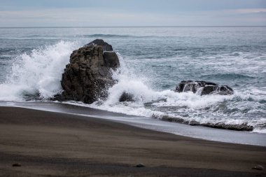 Kara bir kumsaldaki karanlık volkanik kayaların sahili dalgalar kıyıya çarpıp püskürürken bulutlu bir gökyüzünün altında sakin bir ufuk çiziyor..