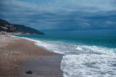 Turkuaz dalgaların kara bulutlu bir gökyüzü altında çarpıştığı bir çakıl taşı sahili manzarası, yamaçta yuva yapmış bir sahil kasabası.