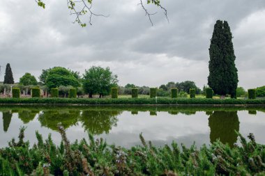 Huzurlu bir yansıma göleti, bulutlu bir gökyüzünün altında tarihi bir İtalyan bahçesinde manikürlü çalılar ve selvi ağaçları yansıtır.