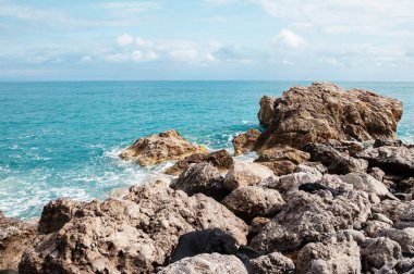 Jagged kayalar parçalı bulutlu bir gökyüzünün altında canlı turkuaz okyanusla buluşur ve sakin bir kıyı manzarası yaratır.