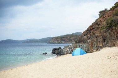 Küçük mavi bir çadır sahilde tek başına bir kaya ve ormanın yanında duruyor.