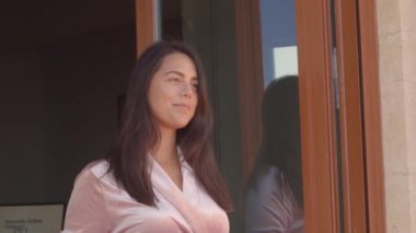 Pembe kadife bornozlu güzel bir kadın, evinin penceresinde duruyor ve güneşli bir sabahta ilk kahvesini içiyor. Görüntü kalitesi yüksek 4K görüntü.