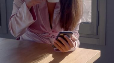 Pembe bornozlu, elinde telefon olan, güneşli bir sabahta kahve içen beyaz bir kız. Görüntü kalitesi hala 4K..