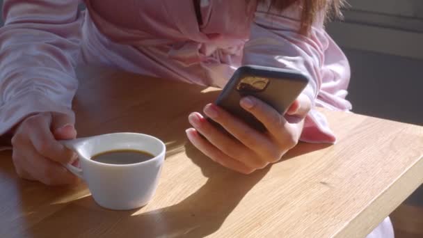 Da vicino, senza volto. Ragazza bianca in accappatoio rosa, con un telefono in mano, bere una tazza di caffè al mattino di sole. Video pinzato 4k di alta qualità. — Video Stock