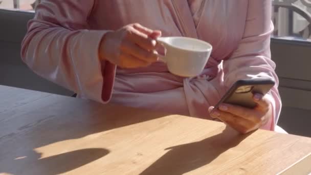 Biała dziewczyna w różowym szlafroku, z telefonem w dłoni, pijąca pierwszą filiżankę kawy w słoneczny poranek. Panele wideo wysokiej jakości 4k materiału. — Wideo stockowe