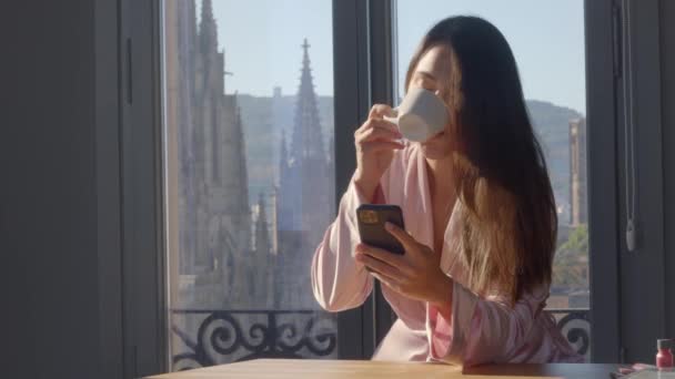 Jovem mista de raça feminina em um robe de seda rosa, com um smartphone na mão está bebendo sua primeira xícara de café com vistas Barcelona sobre o fundo. Ainda vídeo de alta qualidade 4k imagens. — Vídeo de Stock