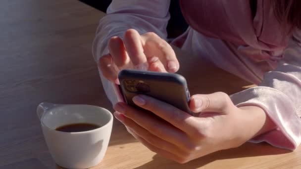 Närbild, inget ansikte. Vit tjej i en rosa morgonrock, med en telefon i handen, dricker vid en kopp kaffe på den soliga morgonen. Konserverad video av hög kvalitet 4k film. — Stockvideo