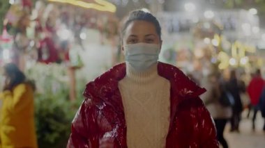 Tıbbi maske takan genç bir kadın kırmızı kürk manto içinde rahat duruyor. Arka planda neşeli bulanık noel ışıkları var. Ortalama yakın çekim 4k video yüksek kaliteli görüntü.