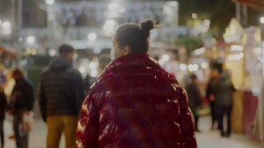 Kırmızı ceketli ve tıbbi maskeli genç, siyah saçlı bayan Noel pazarında dolaşıyor. Arka planda bulanık dekorasyon. 4k 'lık yüksek kaliteli video görüntüsü.