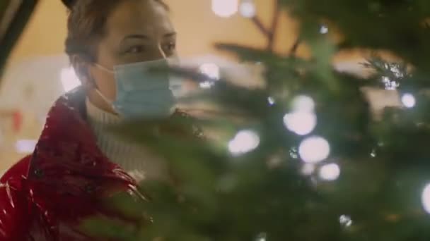 Jeune femme aux cheveux bruns dans un masque médical derrière un sapin de Noël regardant autour. Décoration floue sur le fond. Vidéo 4k pané séquences de haute qualité. — Video