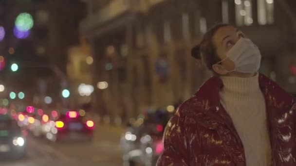 Donna caucasica con una maschera di protezione medica che si guarda intorno. Centro città notturno, stagione festiva. Panning medio primo piano, 4K riprese video di alta qualità. — Video Stock