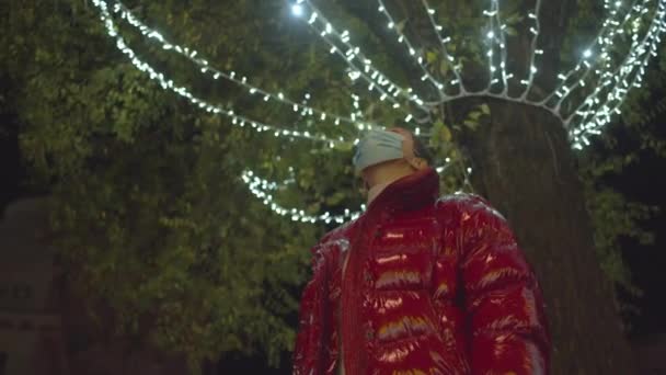 A young caucasian woman in a surgical mask watching up on the street festive Christmas lights. 4k video high quality footage. — Stock Video