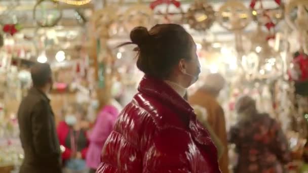 Νεαρή γυναίκα με μάσκα ιατρικού προσώπου τριγυρνάει στην Χριστουγεννιάτικη αγορά. Μέτριο κοντινό πλάνο 4k βίντεο υψηλής ποιότητας. — Αρχείο Βίντεο
