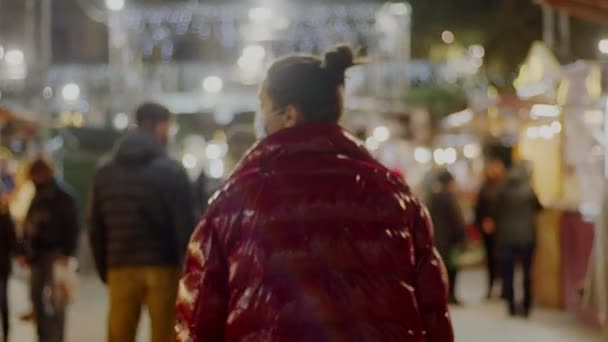 Jovem senhora de cabelo escuro em um casaco vermelho e máscara facial médica andando pelo mercado de Natal. Decoração borrada no fundo. Panned 4k vídeo de alta qualidade de imagens. — Vídeo de Stock