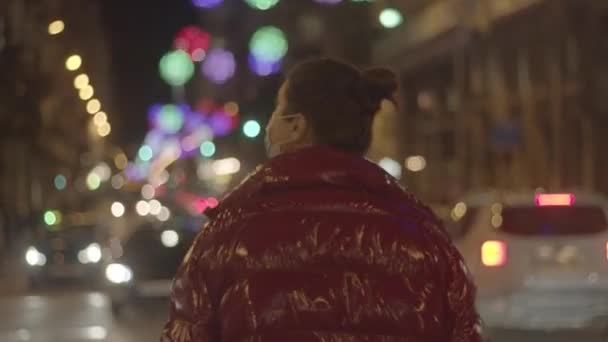 COVID-19 마스크를 쓴 젊은 여성이 겨울 휴가 도시의 밤을 즐기고 있습니다. 교통 체증이 심한 중앙 도로. 미디엄 클로즈업 , 4K 고품질 비디오 필름. — 비디오