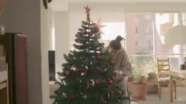 Koyu renk saçlı ırk kızı dekorasyonu ve evde yapay Noel ağacı. Orta Geniş Kamera 4K yüksek kaliteli video görüntüsü.