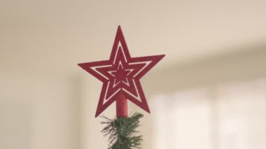 Güneşli evdeki bir Noel ağacının tepesinde parlak kırmızı bir yıldız süslemesi. Kapalı 4K yüksek kaliteli video.