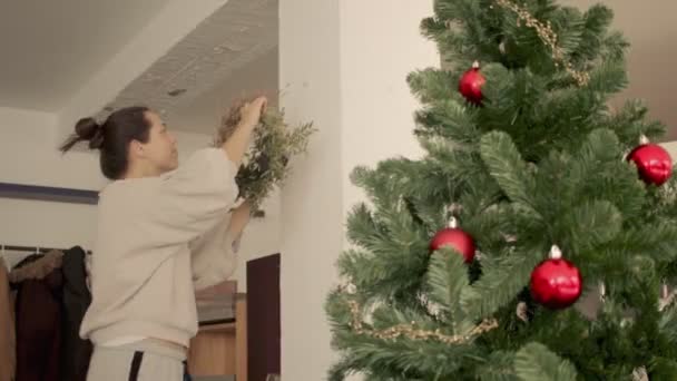 Mujer joven caucásica con el pelo oscuro en un moño está poniendo una corona de Navidad en la pared. Vista lateral Cowboy disparo 4k video de alta calidad. — Vídeo de stock