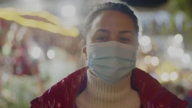 Tıbbi maske takmış genç bir esmer kırmızı kürk manto içinde rahat rahat izliyor. Arka planda neşeli bulanık noel ışıkları var. 4k 'lık yakın çekim yüksek kaliteli görüntü.