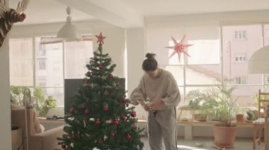 Koyu saçlı, beyaz bir kız evde yapay bir Noel ağacını çelenkle süslüyor. Orta Geniş Kamera 4K yüksek kaliteli video görüntüsü.