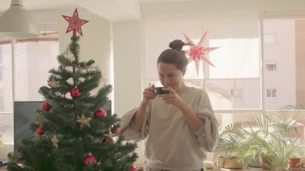 Chica de raza mixta de pelo oscuro bebiendo café y viendo el árbol de Navidad en casa. Cámara de tamaño mediano 4k filmación de vídeo de alta calidad. — Vídeos de Stock