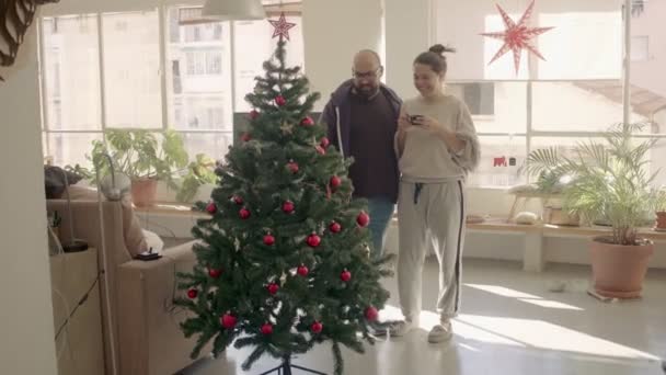 人工的なクリスマスツリー上の装飾を見てホームウェアのカップル。フルショット高品質4kビデオ映像. — ストック動画