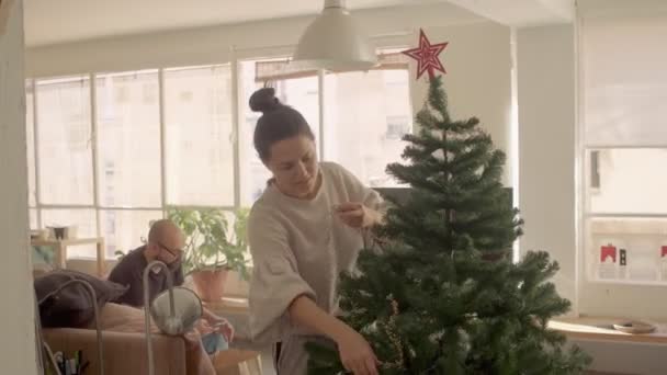Mörkt hår blandras flicka gör upp och konstgjord julgran. En man med telefonen i bakgrunden. Medelhög bild 4k hög kvalitet videofilmer. — Stockvideo