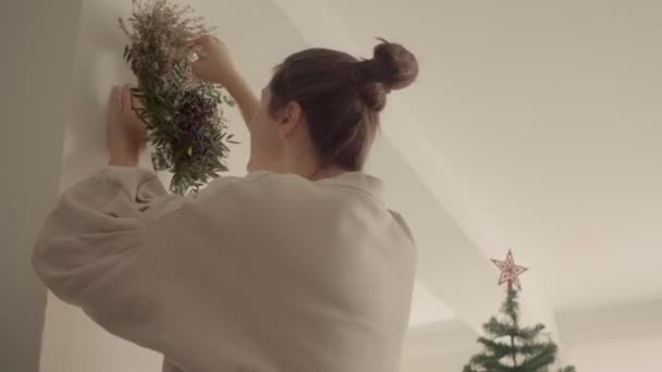 Blank meisje met donker haar in een broodje hangt een kerstkrans op de muur. Omhoog bekijken medium shot 4k hoge kwaliteit video. — Stockvideo