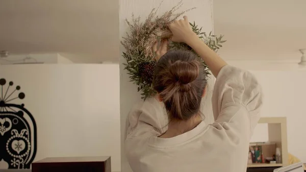 Menina caucasiana com cabelo escuro em um pão está colocando uma coroa de Natal na parede. Up vista médio tiro de alta qualidade foto jpg. — Fotografia de Stock