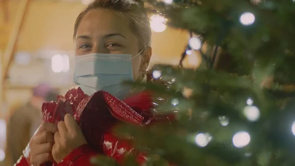 Jovem de cabelo escuro com jaqueta vermelha e máscara facial médica olhando por trás de uma árvore de Natal. Retrato médio de close-up. — Fotografia de Stock
