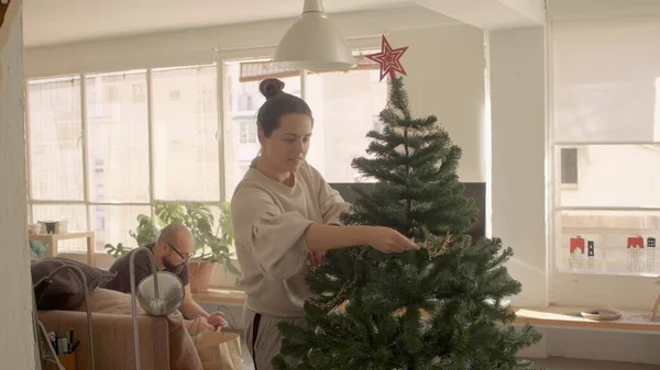 Koyu renk saçlı, melez kadın dekorasyonu ve evde yapay Noel ağacı. Kanepede oturan adam. Orta Geniş Kamera yüksek çözünürlüklü jpg görüntüsü — Stok fotoğraf