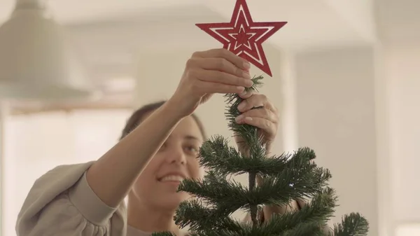 Brunetka biała młoda kobieta umieszcza czerwoną gwiazdę na sztucznej choince. Średni strzał wysokiej jakości zdjęcie jpg. Obrazek Stockowy