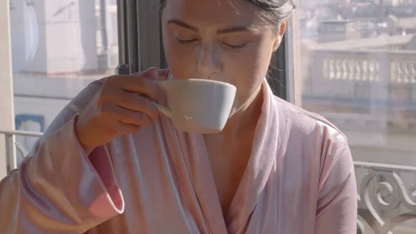 Kaukázusi fiatal nő rózsaszín köntösben issza az első csésze kávét a napsütéses reggelen. Közepes közelkép nagyfelbontású jpg kép — Stock Fotó