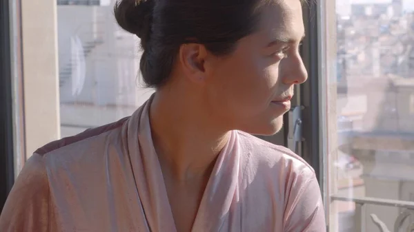 Menina caucasiana em um robe de veludo rosa, olhando para fora da janela na manhã ensolarada. Retrato de close-up imagem jpg de alta resolução — Fotografia de Stock