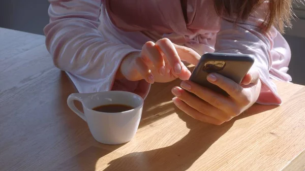 Ragazza bianca in accappatoio rosa, con uno smartphone in mano e una tazza di caffè sul tavolo al mattino di sole. Chiudi nessuna immagine jpg ad alta risoluzione — Foto Stock