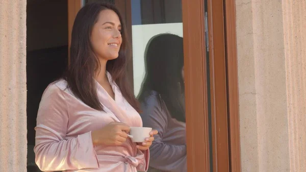 Attractive caucasian young woman in a rose velvet robe with a ceramic cup standing near the open window with a cup of coffee in the sunny morning, smiling. High-quality medium shot portrait Telifsiz Stok Imajlar